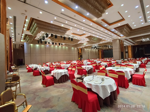 Großer Ballsaal mit weiß gedeckten Tischen, Gläsern, Tellern, Besteck und Dekoration an den Wänden sowie beleuchteter Decke, mit einem Wasserzeichen unten rechts.