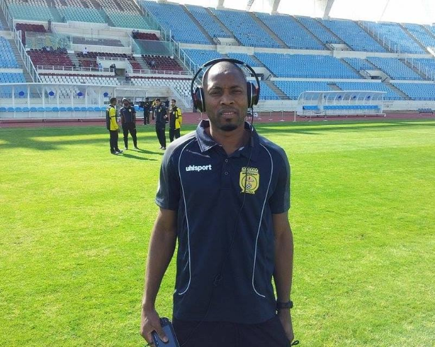 Ein Mann mit Headset, wahrscheinlich der Trainer der ghanaischen Nationalmannschaft, steht auf einem Fussballfeld in der Nähe eines Stadions und hält ein Objekt, während im Hintergrund Menschen zu sehen sind und der Himmel über ihnen ist.