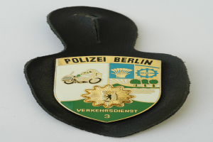 Ein Polizeidienstabzeichen mit der Aufschrift "Polizei Berlin" in fetter schwarzer Schrift auf einem blauen Schild mit einem weißen Stern und einem weißen Rand.