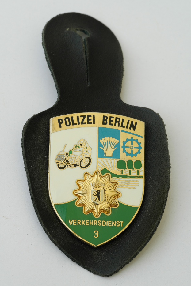 Ein Polizeidienstabzeichen mit der Aufschrift "Polizei Berlin" in fetter schwarzer Schrift auf einem blauen Schild mit einem weißen Stern und einem weißen Rand.