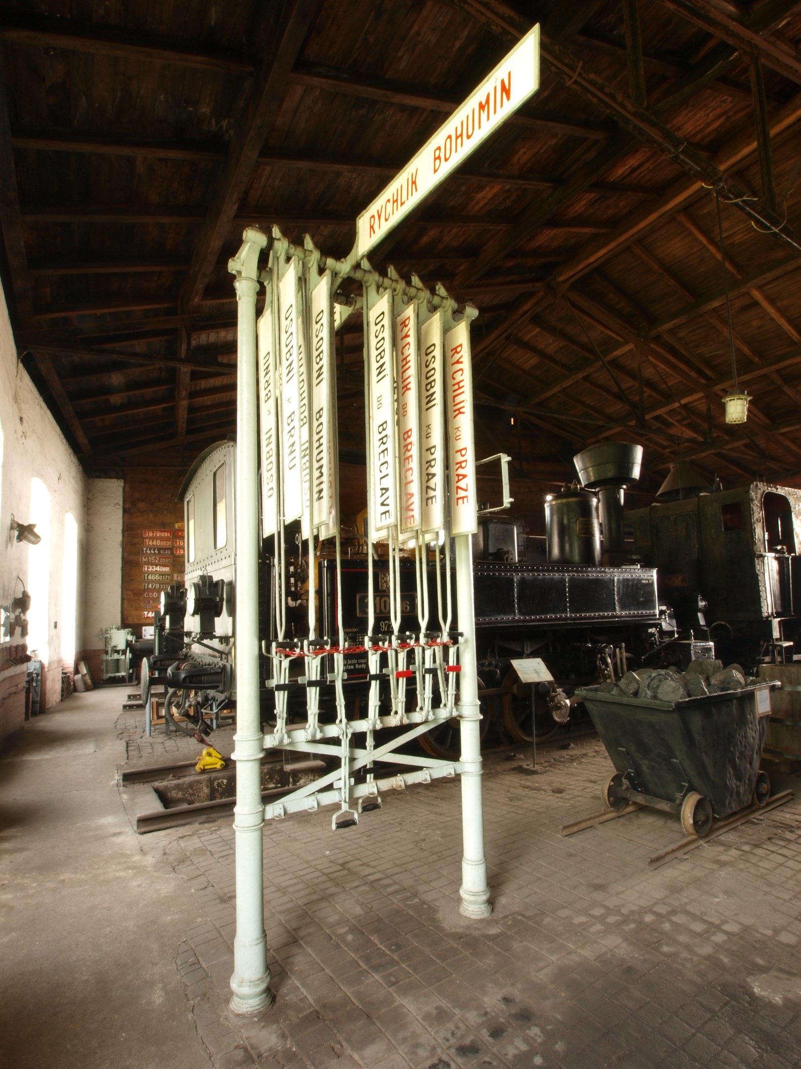 Ein Bahnsteig mit einem Zug auf den Gleisen, ein Bodenwagen, ein an einem Pfahl befestigtes Schild, eine Wand links, eine Decke oben und eine zentrale Signalbox, die die Ankunft des Zuges steuert.