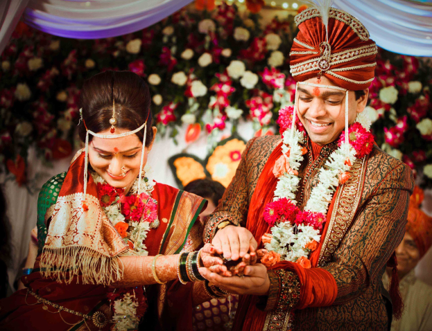 Two people in traditional Indian wedding attire exchange rings as others observe in a decorated setting with flowers and a curtain backdrop.