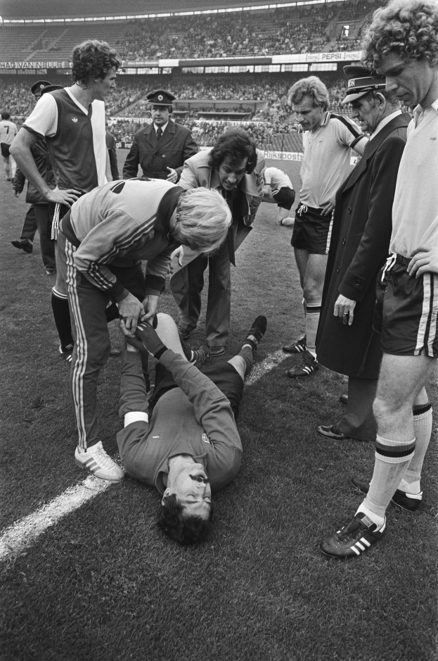 Schwarzes und weißes Bild eines Fußballspielers, der auf dem Boden liegt und von einer Gruppe Männer umgeben ist, mit einer Menge im Stadionhintergrund.