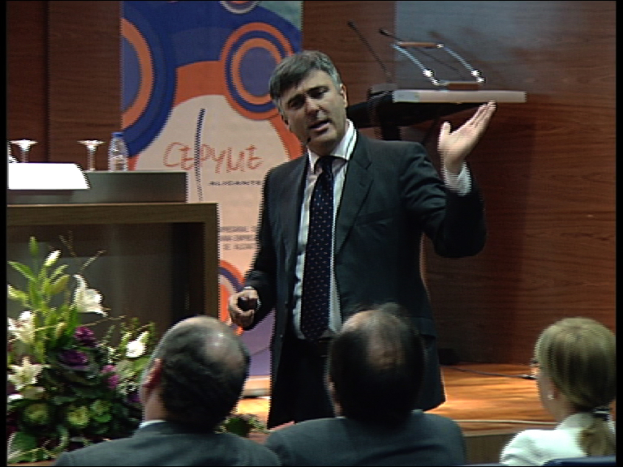 Ein Mann in einem schwarzen Blazer steht an einem Podium mit Mikrofonen und spricht in ein tragbares Mikrofon. In der Nähe befindet sich ein Blumentopf oder ein Blumenstrauß. Vor ihm sitzen zwei Männer und eine Frau auf Stühlen. Hinter ihm steht ein Tisch mit einem Buch, einer Wasserflasche und anderen Gegenständen. Im Hintergrund ist eine braune Wand und eine farbige Tafel mit Text zu sehen.