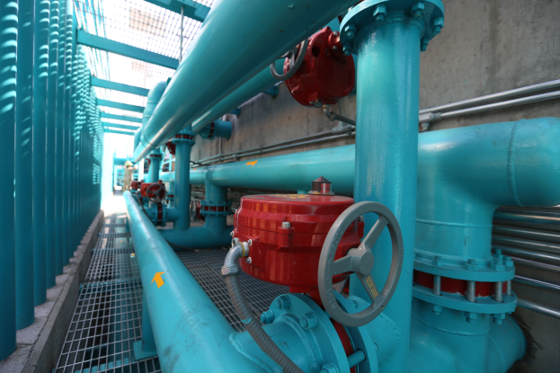 Eine große Gruppe von blauen Rohren und Ventilen in einem Gebäude, mit einem prominenten roten Ventil in der Mitte, Metallstangen links und einer Wand im Hintergrund, die wahrscheinlich den Prozess des Baus einer Gasleitung darstellt.