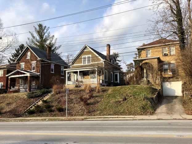 Eine Reihe von Häusern entlang einer Straße mit Fenstern, Türen, Säulen, Geländern, Stufen, Pflanzen, Gras, Bäumen und einem Pfahl mit einer Tafel unter einem bewölkten Himmel, repräsentiert eines der teuersten Häuser zum Verkauf in Detroit.