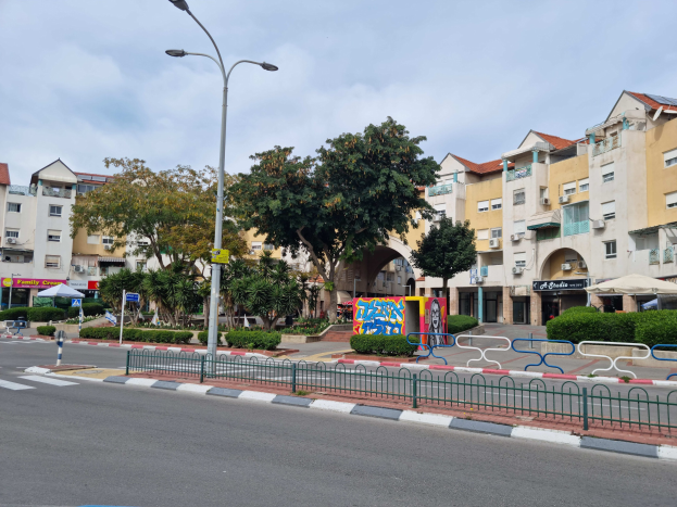 A city street in Penang, Malaysia, with tall buildings in the background, street furniture, greenery, and signage under a cloudy sky.