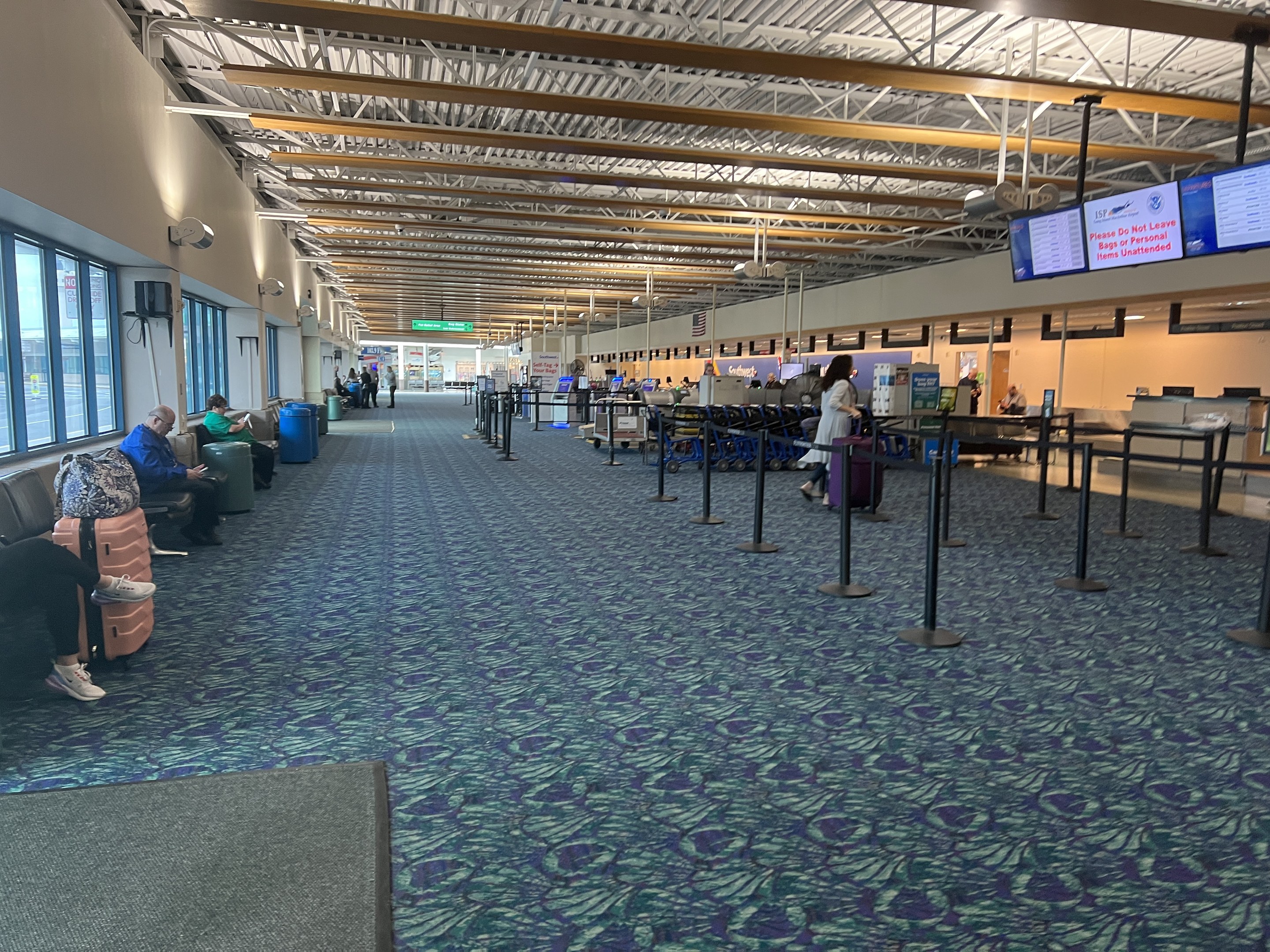 Lange Schlange von Menschen in einem Flughafen-Terminal mit Sitzplätzen, stehenden Passagieren, Wandbildschirmen, Deckenleuchten, Glasfenstern auf der linken Seite und Informationsanzeigen im Hintergrund, die mit "Vereinigte Staaten von Amerika (USDA)" beschriftet sind.