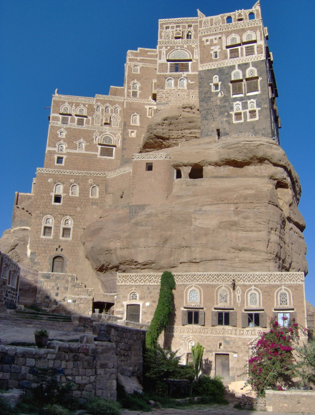 Ein Schloss mit Fenstern und Türen, umgeben von Pflanzen, unter einem blauen Himmel.