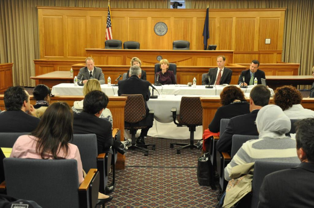 Tagungsraum mit einer Gruppe von Menschen, die auf Stühlen sitzen, zwei ausländische Flaggen, ein oranger Schreibtisch mit Wasserflaschen und ein Mikrofon.