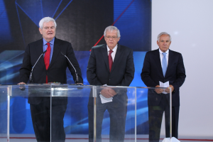 Ein älterer Mann in einem schwarzen Mantel und roter Krawatte steht an einem transparenten Rednerpult auf einer Bühne, mit einer blauen und weißen Fahne im Hintergrund.