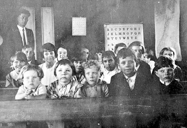 Ein Schwarz-Weiß-Foto einer Klasse mit Kindern auf Bänken im Vordergrund, einer Person im Hintergrund und einer Alphabettafel und einem Kalender an der Wand dahinter.