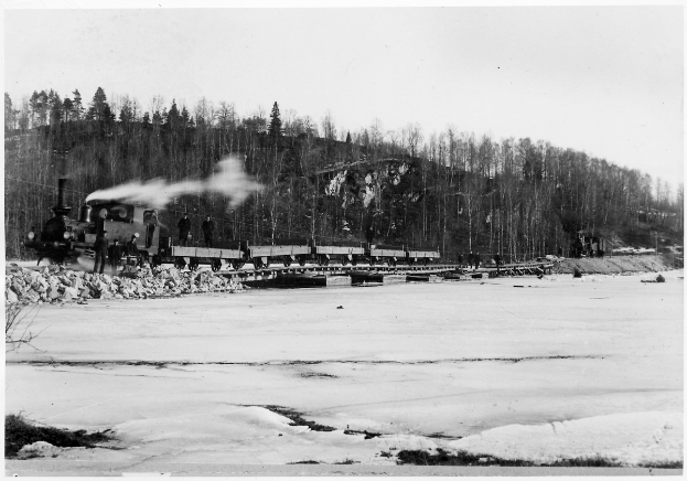 Ein Schwarz-Weiß-Foto eines Zuges, der über einen zugefrorenen Fluss fährt, mit Menschen auf einer Brücke, Bäumen, einem Hügel und dem Himmel im Hintergrund.