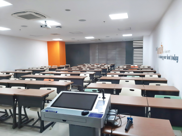 A classroom with desks and chairs in rows, a ceiling-mounted projector, and objects on tables, featuring text on the right wall indicating a private school in Singapore.