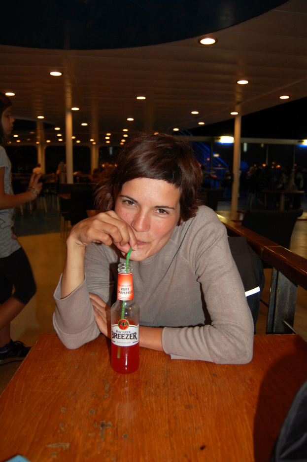 Eine Frau sitzt auf einem Stuhl an einem Holztisch, trinkt ein Bier, mit einer Person, die links von ihr steht, unter einem Dach mit Beleuchtungskörpern.