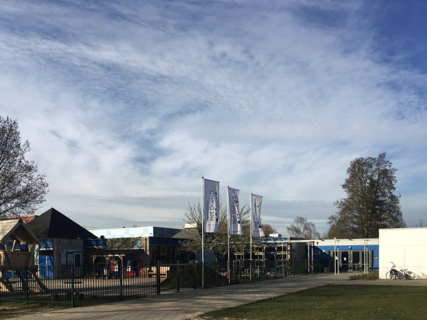 Großes Schulgebäude mit Metallzaun, Rasenfläche, Weg, Bäumen, Bannern, einem geparkten Fahrrad, nahen Häusern und einem bewölkten Himmel, das als neuer Spielplatz für Kinder bezeichnet ist.
