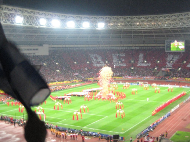 Ein Stadion mit Flutlicht, Eisengittern, Zuschauern, stehenden Personen, Werbetafeln, Anzeigetafeln, Überwachungskamera und Fahnen und Dekorationen.