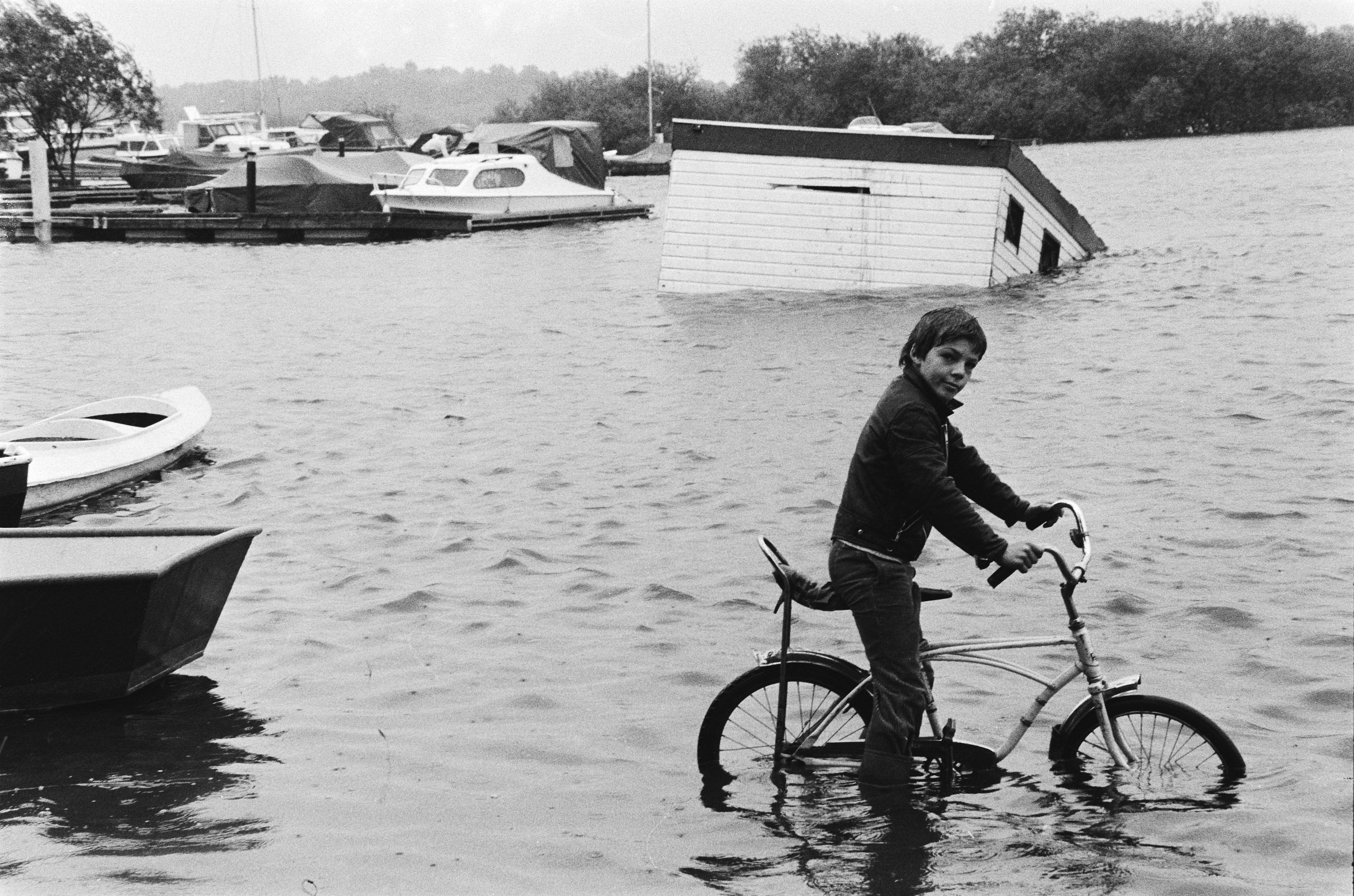 Ein junger Junge fährt mit einem Fahrrad durch eine überflutete Straße, mit Booten links, einem Haus in der Mitte und Bäumen und Himmel im Hintergrund, dargestellt in Schwarz-Weiß.