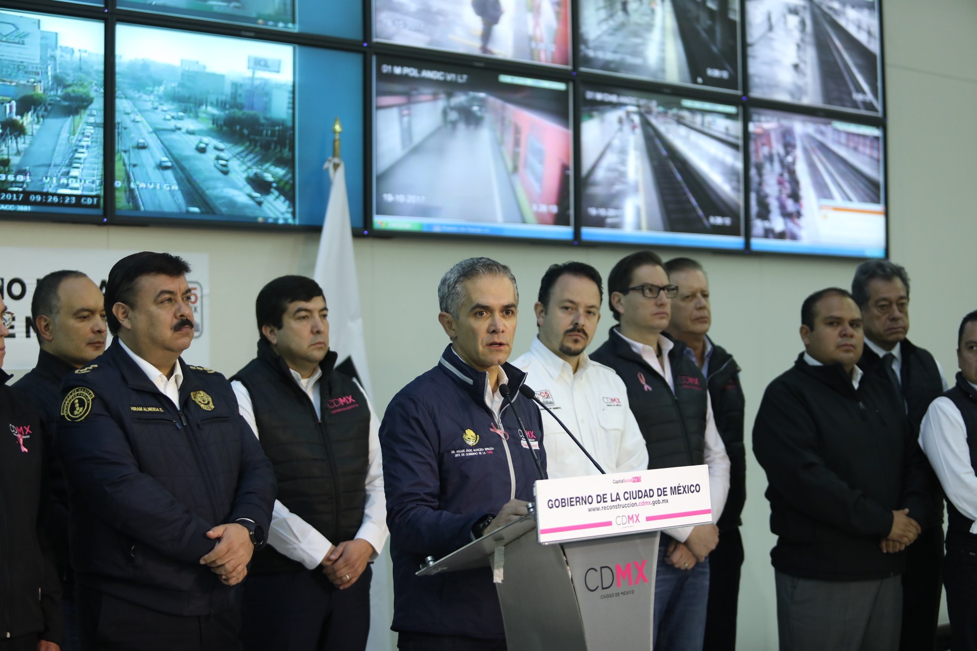 Gruppe von Männern vor einem Rednerpult mit einem Mikrofon und einer Tafel mit der Aufschrift "Mexikanische Zoll- und Grenzschutzbehörde", mit Fernsehgeräten und einer Flagge an der Wand dahinter.