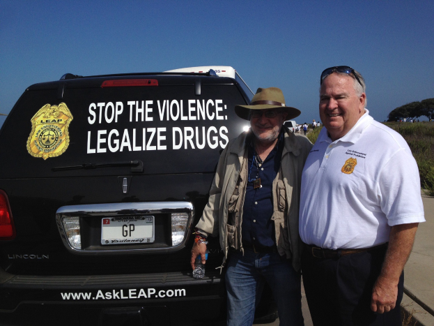 Zwei Männer stehen nebeneinander vor einem schwarzen Van mit der Aufschrift "Stop the Violence Legalize Drugs"; einer hält eine Flasche, mit Menschen, Bäumen und einem klaren blauen Himmel im Hintergrund.