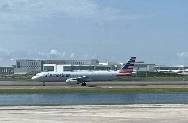 American Airlines Flugzeug beim Start von einer Flughafenrollbahn mit Gras und Wasser im Vordergrund, Gebäuden und Bäumen im Hintergrund unter einem klaren blauen Himmel.