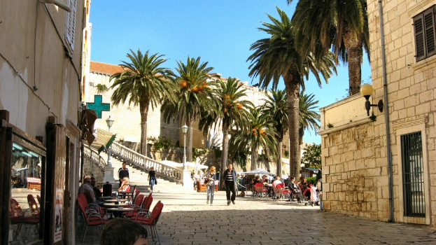 Zwei Paare spazieren auf einem Fußweg, der von offenen Restaurants mit Tischen und Stühlen gesäumt ist, Kokospalmen im Hintergrund und eine Backsteinmauer mit Fensterrost und Tür auf der rechten Seite.