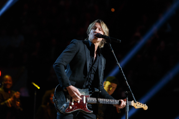 Keith Urban performing on stage with a guitar, a microphone, and a band in a dimly lit venue.