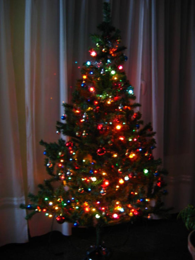 Ein Weihnachtsbaum mit Lichterschmuck steht vor einem Vorhang und einem Topf.