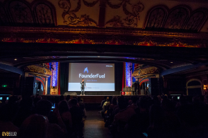 Eine Person steht auf einer Bühne mit einem Bildschirm im Hintergrund, schwarzen Vorhängen und einem Publikum auf Stühlen sitzend.