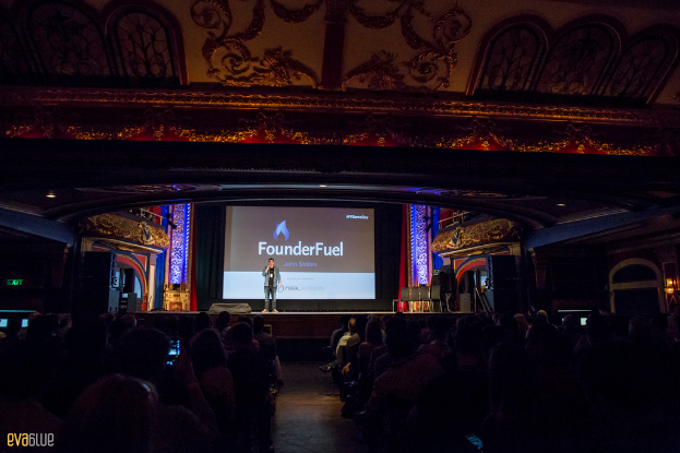 Eine Person steht auf einer Bühne mit einem Bildschirm im Hintergrund, schwarzen Vorhängen und einem Publikum auf Stühlen sitzend.
