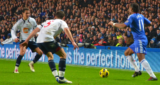 Personen mit einem Ball im Vordergrund, eine Tafel mit Text in der Mitte, Kameras hinter der Tafel und eine Gruppe von Personen im Hintergrund.