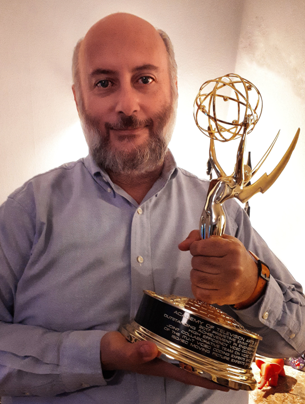 Ein Mann lächelt, während er einen Emmy Award hält, mit verschiedenen Gegenständen auf dem Tisch vor ihm und einer Wand im Hintergrund.