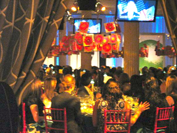 Eine gruppe menschen sitzt an tischen mit essen, fernsehern, lichtern, hängenden papieren und vorhängen in einem raum, der wie eine party aussieht.