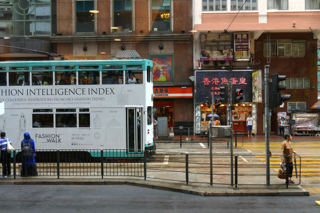 Ein Doppeldeckerbus fährt auf einer von hohen Gebäuden gesäumten Straße, mit Fußgängern auf dem Gehweg und Verkehrsampeln im Hintergrund.