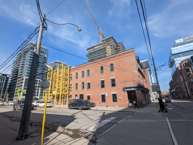 Eine belebte Stadtstraße mit fahrenden Fahrzeugen, Fußgängern auf dem Gehweg, Strommasten mit Drähten und Gebäuden, darunter ein teilweise errichteter Hochhaus mit einem Kran im bewölkten Himmel.