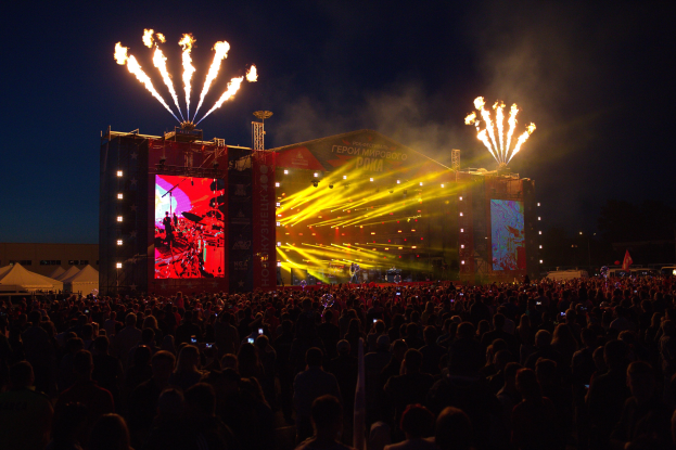 Eine große Menschenmenge auf einem Musikfestival mit einer Bühne, auf der Künstler unter hellen Scheinwerfern spielen, Banner, Zelte links und ein dunkler Himmel mit Feuerwerk.