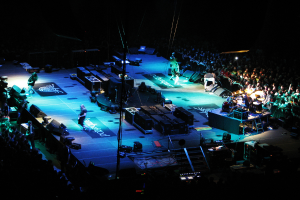 Menschen, die auf einer Bühne mit Boxen, Lautsprechern, Lampen und Mikrofonen performen, mit einer großen Menge auf beiden Seiten und einem dunklen Hintergrund.