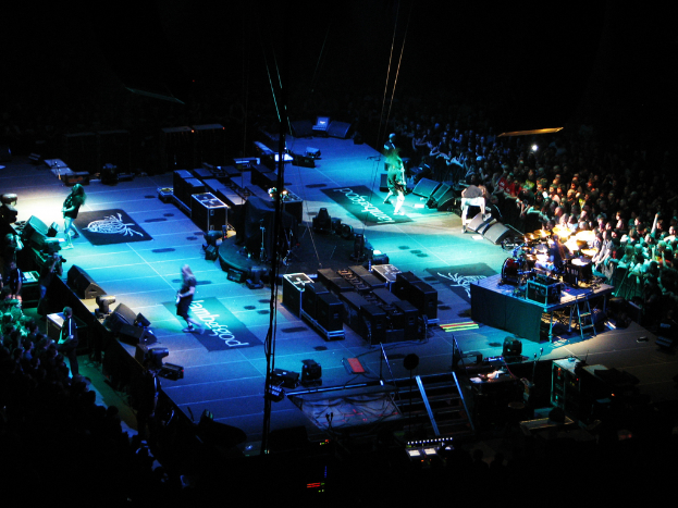 Menschen, die auf einer Bühne mit Boxen, Lautsprechern, Lampen und Mikrofonen performen, mit einer großen Menge auf beiden Seiten und einem dunklen Hintergrund.