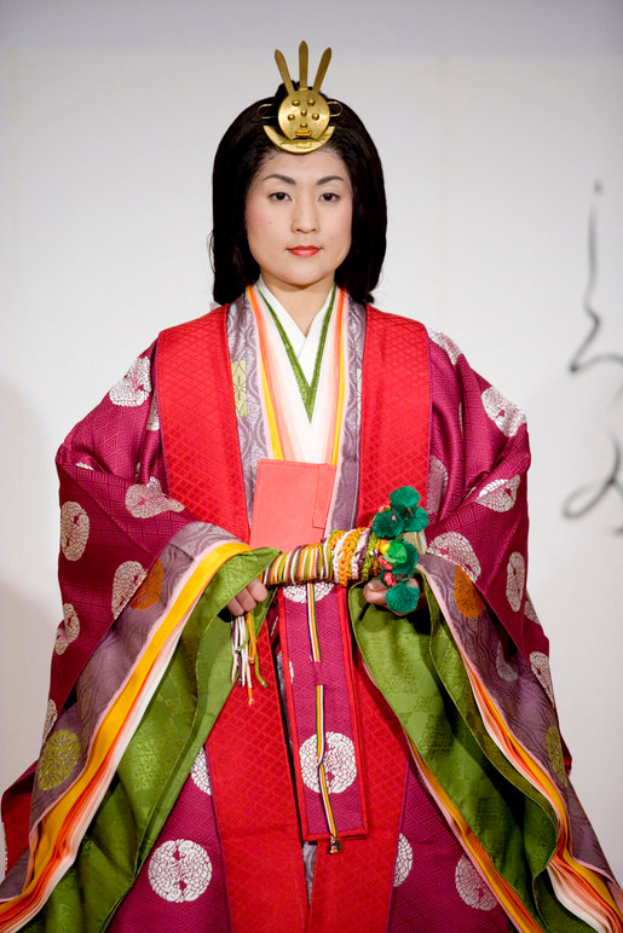 Eine Frau in einem traditionellen japanischen Kimono, das ein leuchtendes rotes und grünes Kleid mit kunstvollen Mustern trägt, steht selbstbewusst auf einem Laufsteg, ihr Haar zu einem ordentlichen Knoten gebunden.