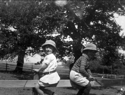 Zwei junge Jungs in Hüten mit Baseballschlägern in einem Park mit Bäumen, einem Zaun und Gras, in Schwarz-Weiß.