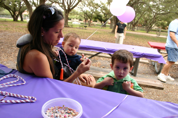 Eine Frau in Schutzbrille hält ein Baby, daneben ein Junge in einem grünen Kleid, neben Tischen mit einer Geigenpartitur und einer Schale mit Würfeln, Ballons und einem Mann rechts, mit Bäumen im Hintergrund.