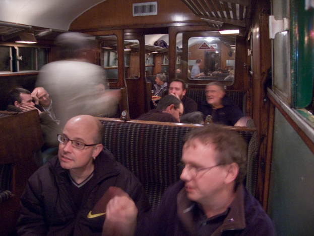 Zwei Männer sitzen auf einem Sitz in einem Abteil, mit einer Gruppe von Menschen, die dahinter sitzen; ein Eingang zwischen Abteilen ist oben sichtbar, und ein Fenster ist auf der rechten Seite.