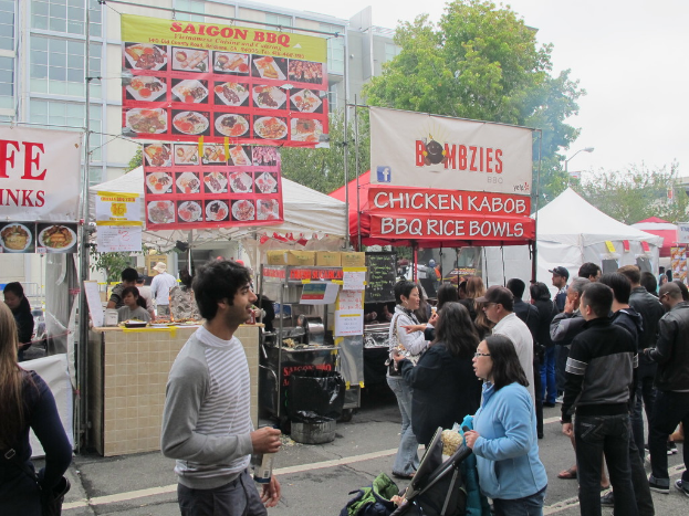 Menschen stehen auf einer Straße vor der Stadt, ein Mann in einem grauen und weißen T-Shirt hält eine Wasserflasche und eine Frau in einer blauen Jacke schiebt einen Kinderwagen. Im Hintergrund sind Stände unter weißen und roten Zelten, Banner mit Text und Gebäude, Bäume und eine Straßenlaterne zu sehen.