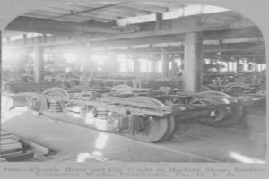 Ein Schwarz-Weiß-Foto eines Elektromotors und eines Lastwagens in einer Fabrik, umgeben von Maschinen, Säulen und einem Dach, mit Text unten.