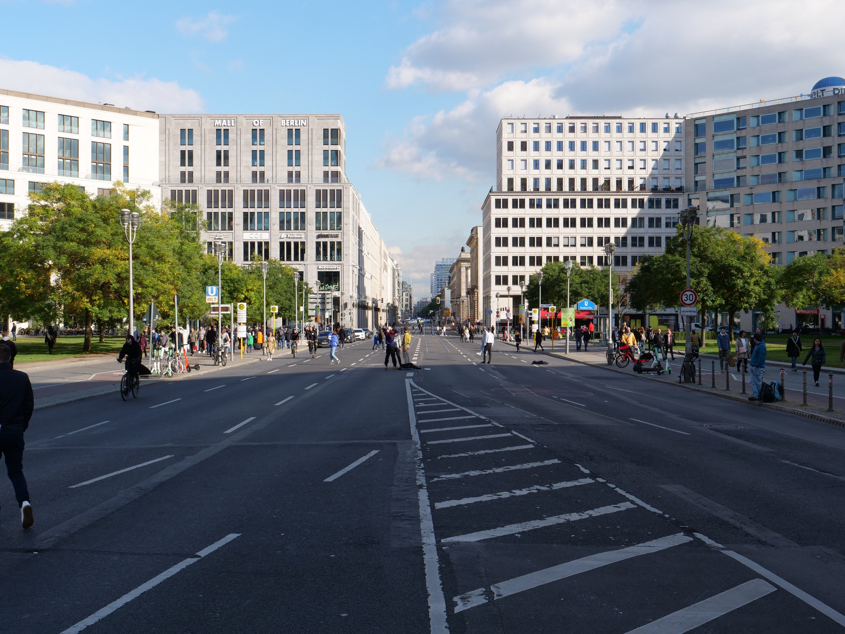 Eine belebte Stadtstraße in Berlin, Deutschland, mit Fußgängern und Radfahrern auf der Straße, hohen Gebäuden, Bäumen, Laternenmasten und Schildern unter einem bewölkten Himmel.