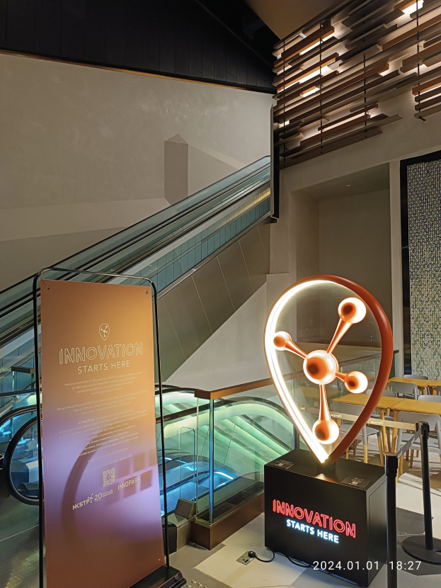 Großes Schild in einem Gebäude mit der Aufschrift "Innovation beginnt hier" und einer Rolltreppe im Hintergrund, Tische und Stühle auf der rechten Seite und eine Tafel auf der linken Seite, die auf die HSTP20 Expo hinweist.