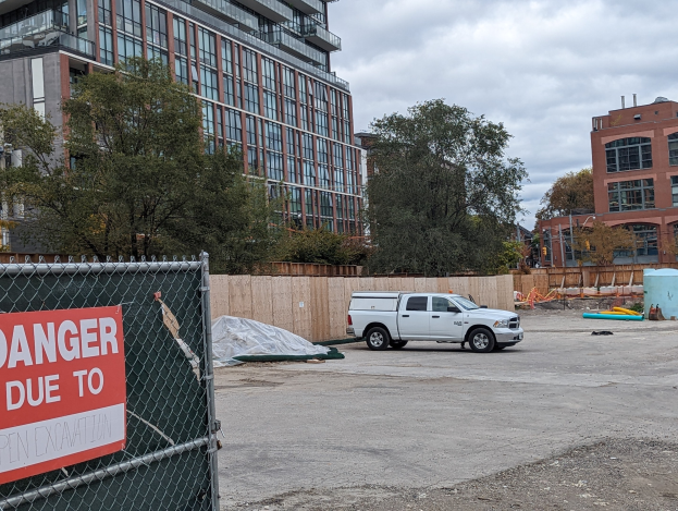 Lastwagen vor einem Gebäude mit einer 'Gefahr wegen offener Baustelle'-Tafel geparkt, Bäume und Gebäude im Hintergrund unter einem klaren blauen Himmel.