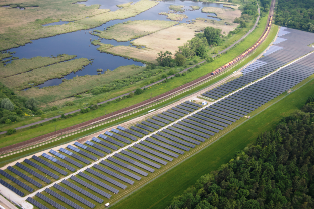 Eine Luftaufnahme einer Solar-Farm mit Panelen auf einem Feld, umgeben von Bäumen, Gras, Wasser und einer nahen Bahnlinie.
