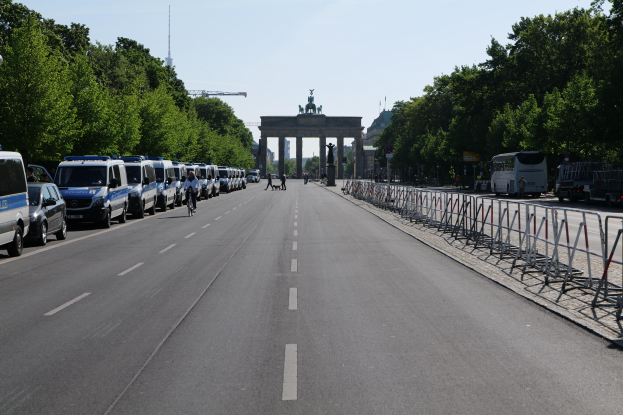 Lange Reihe von Polizeiwagen am Straßenrand vor dem Brandenburger Tor geparkt, mit Menschen auf Fahrrädern und Absperrungen auf der Straße, Bäume an den Seiten und ein Bogen mit Statuen im Hintergrund.