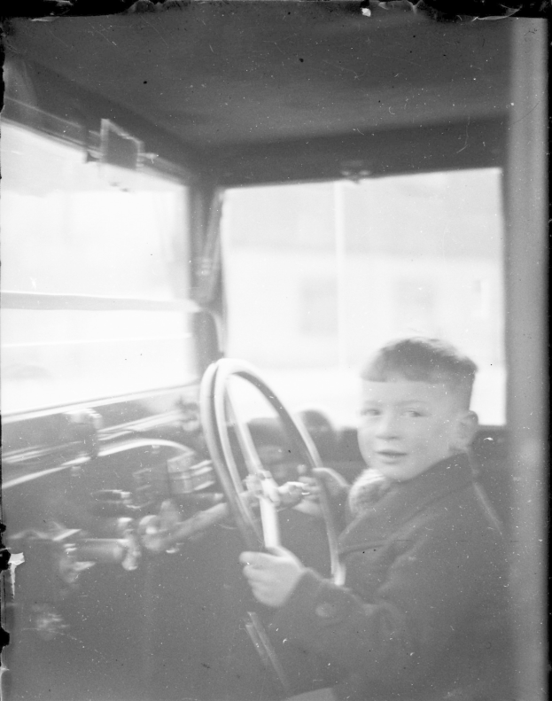 Ein junger Junge in Schwarz-Weiß sitzt im Fahrersitz eines Autos und hält das Lenkrad konzentriert fest.